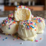 A stack of white chocolate coated sugar cookie truffles decorated with colorful sprinkles, one is bitten to show the soft center.