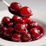 A spoonful of thick, glossy cherry pie filling being lifted from a white bowl filled with vibrant red cherries.