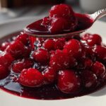 A spoonful of thick, glossy cherry pie filling being lifted from a white bowl filled with vibrant red cherries.