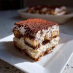 A perfect square slice of homemade tiramisu recipe served on a white plate, topped generously with cocoa powder.