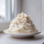 A generous mound of perfectly piped, creamy vanilla buttercream frosting sitting on a small white saucer.