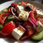Close-up of a vibrant greek salad featuring large chunks of feta cheese, red tomatoes, cucumber, red onion, and Kalamata olives.