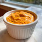 Close-up of bright orange, creamy smash burger sauce speckled with red and black spices in a small white bowl.