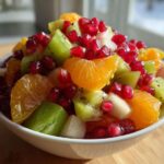 Close-up of a bright winter fruit salad featuring orange segments, green kiwi chunks, and bright red pomegranate seeds in a white bowl.