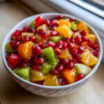 Close-up of a vibrant winter fruit salad featuring bright orange segments, green kiwi chunks, and ruby red pomegranate seeds.