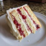 A close-up slice of white chocolate raspberry cake showing three layers of vanilla cake, white frosting, and bright red raspberry filling.