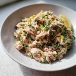 Close-up of a bowl of fresh salmon salad mixed with chopped dill and lemon zest, served with a lemon wedge.