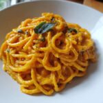 A close-up of spaghetti coated in a rich, orange butternut squash pasta sauce, topped with fresh sage leaves.