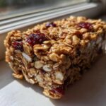A close-up, sunlit view of a thick, chewy no bake granola bar packed with oats and dried cranberries.