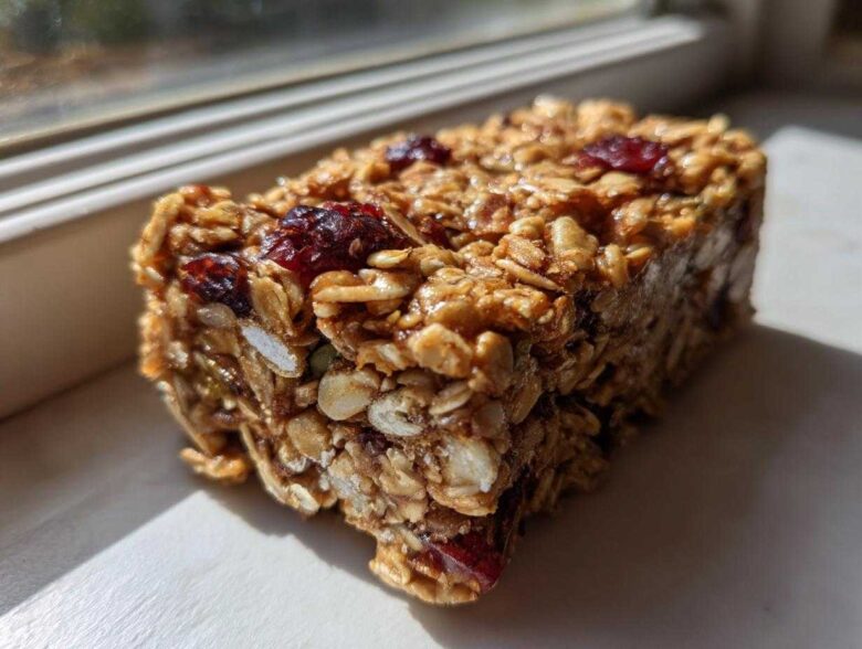 A close-up, sunlit view of a thick, chewy no bake granola bar packed with oats and dried cranberries.