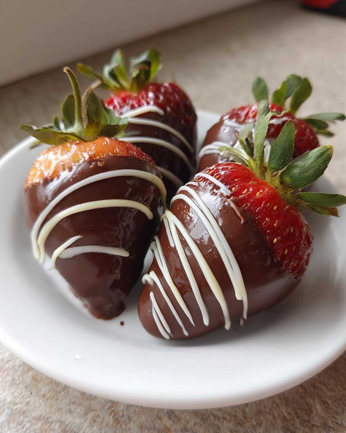 Four fresh chocolate covered strawberries, drizzled with white chocolate, served on a small white plate.