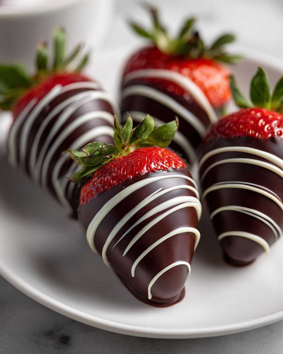 Close-up of four chocolate covered strawberries drizzled with white chocolate on a white plate.