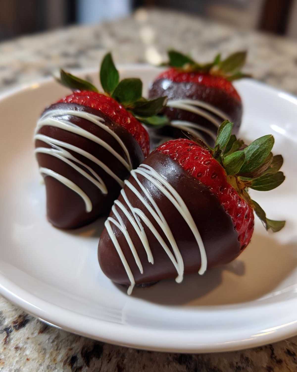 Three freshly made chocolate covered strawberries with a white chocolate drizzle, resting on a white plate.