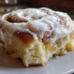 Close-up of a soft cinnamon roll generously topped with thick, glossy Copycat Cinnabon Cream Cheese Frosting on a white plate.