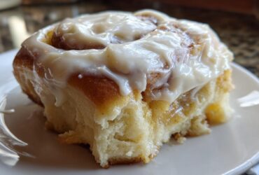 Close-up of a soft cinnamon roll generously topped with thick, glossy Copycat Cinnabon Cream Cheese Frosting on a white plate.