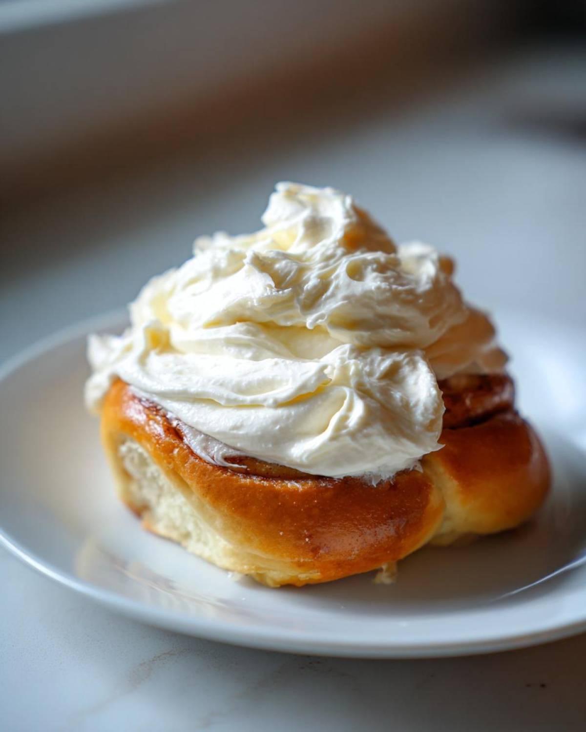 A freshly baked cinnamon roll generously topped with thick, creamy Copycat Cinnabon Cream Cheese Frosting on a white plate.