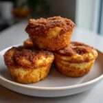 Three golden brown cinnamon sugar french toast muffins stacked on a light gray plate, featuring a thick, sugary topping.