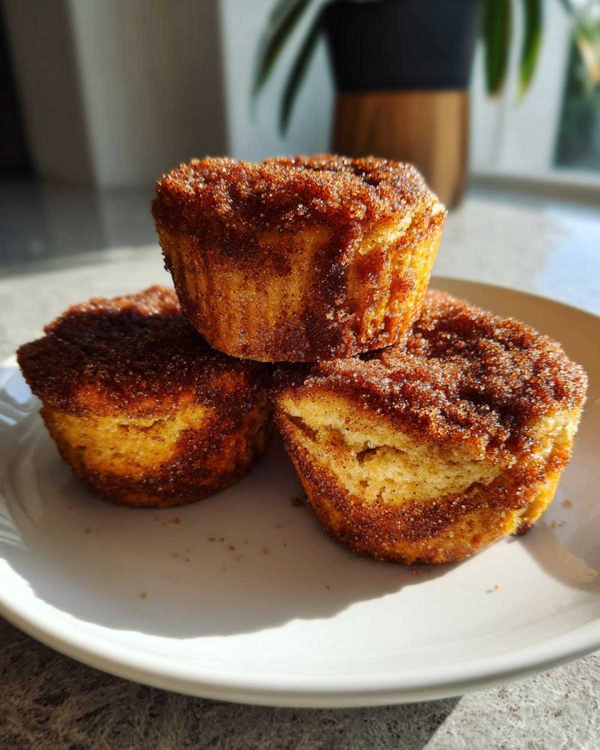 Three golden cinnamon sugar french toast muffins stacked on a white plate, heavily coated in cinnamon sugar.