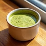A small white bowl filled with vibrant green Copycat Panera Green Goddess Dressing, sitting on a wooden surface.