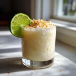 A creamy, blended Iced Pineapple Coconut Mocktail topped with toasted coconut and garnished with a fresh lime wheel.