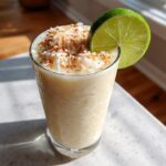 A tall glass of creamy Iced Pineapple Coconut Mocktail topped with toasted coconut flakes and a lime wheel.