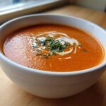 Close-up of a white bowl filled with rich, orange tomato bisque, garnished with cream swirl and fresh herbs.