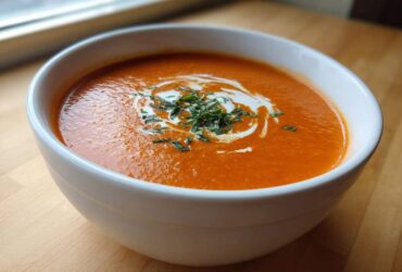 Close-up of a white bowl filled with rich, orange tomato bisque, garnished with cream swirl and fresh herbs.