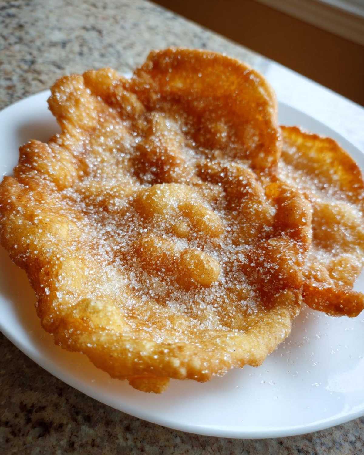 Close-up of two golden, crispy buñuelos heavily dusted with granulated sugar, resting on a white plate.