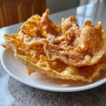 Close-up of crispy, golden brown buñuelos heavily dusted with sparkling sugar served on a small white plate.