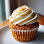 A single Gluten-Free Carrot Cake Cupcake topped with a swirl of cream cheese frosting and orange zest.