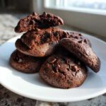 A stack of rich, dark Gluten-Free Chocolate Mint Cookies studded with chocolate chips, displayed on a white plate.