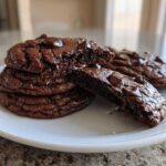 A stack of rich, dark Gluten-Free Chocolate Mint Cookies on a white plate, one cookie broken open to show the fudgy interior and melted chocolate chips.