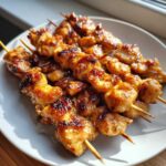 Close-up of several golden-brown, glazed chicken skewers resting on a white plate, showing char marks.
