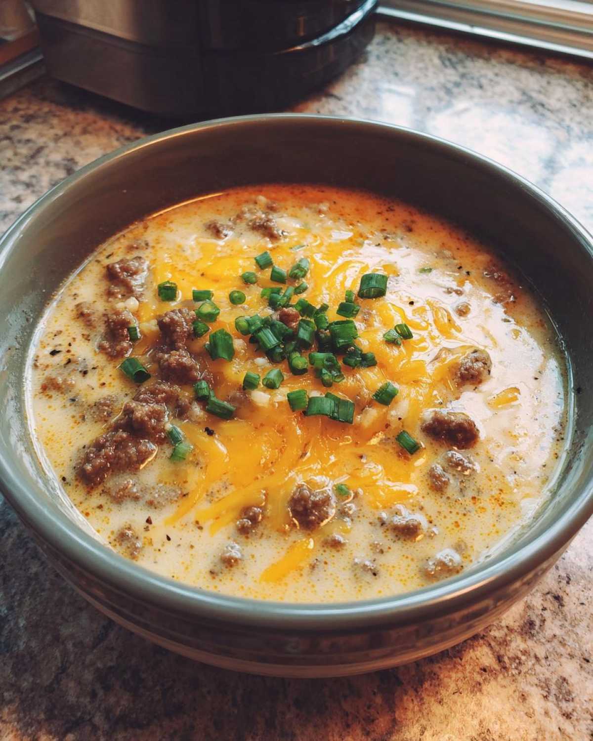 A close-up of a bowl of creamy Keto Cheeseburger Soup topped with melted cheddar cheese and fresh green chives.