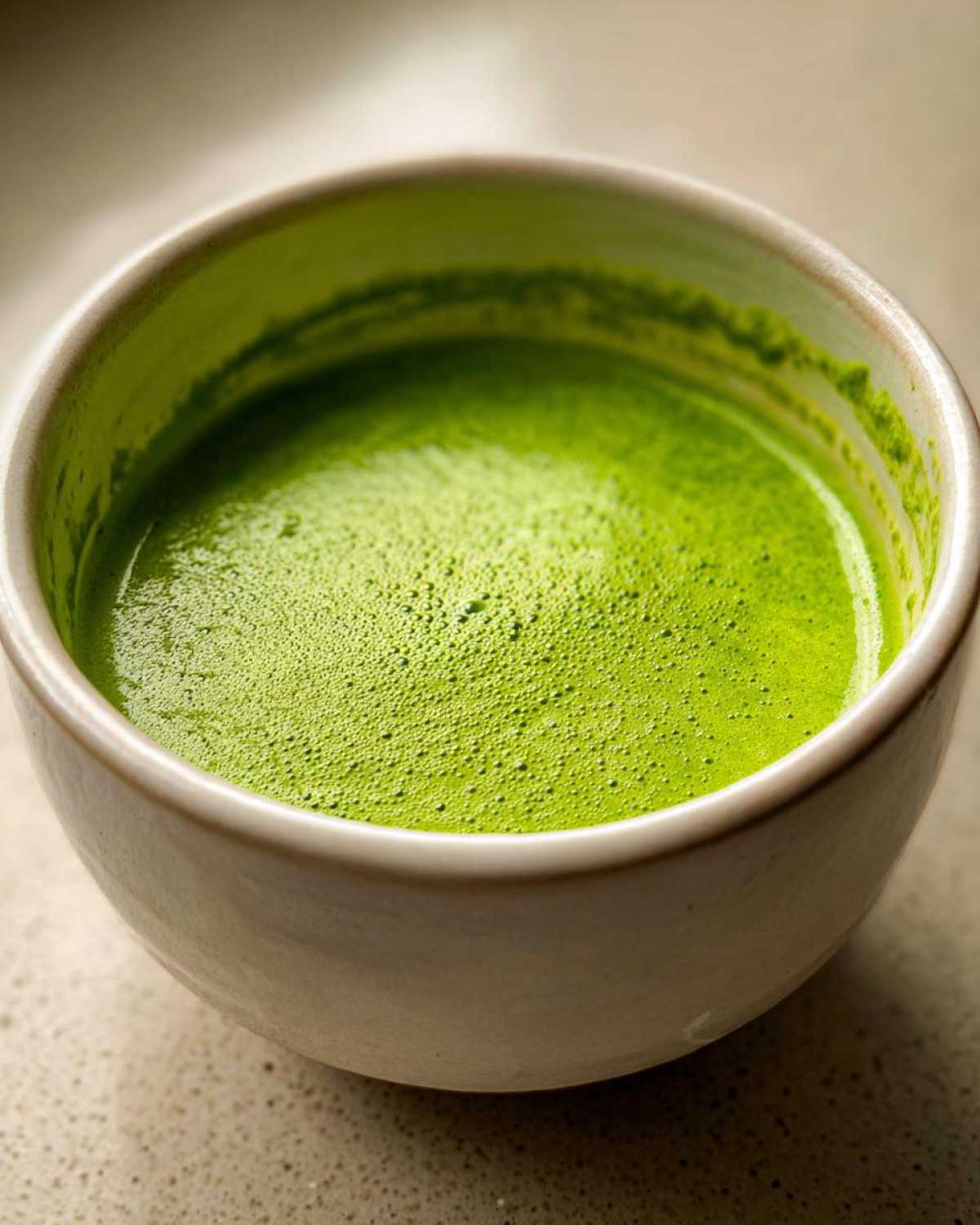 Close-up of vibrant green, frothy liquid, likely matcha, in a light ceramic bowl.