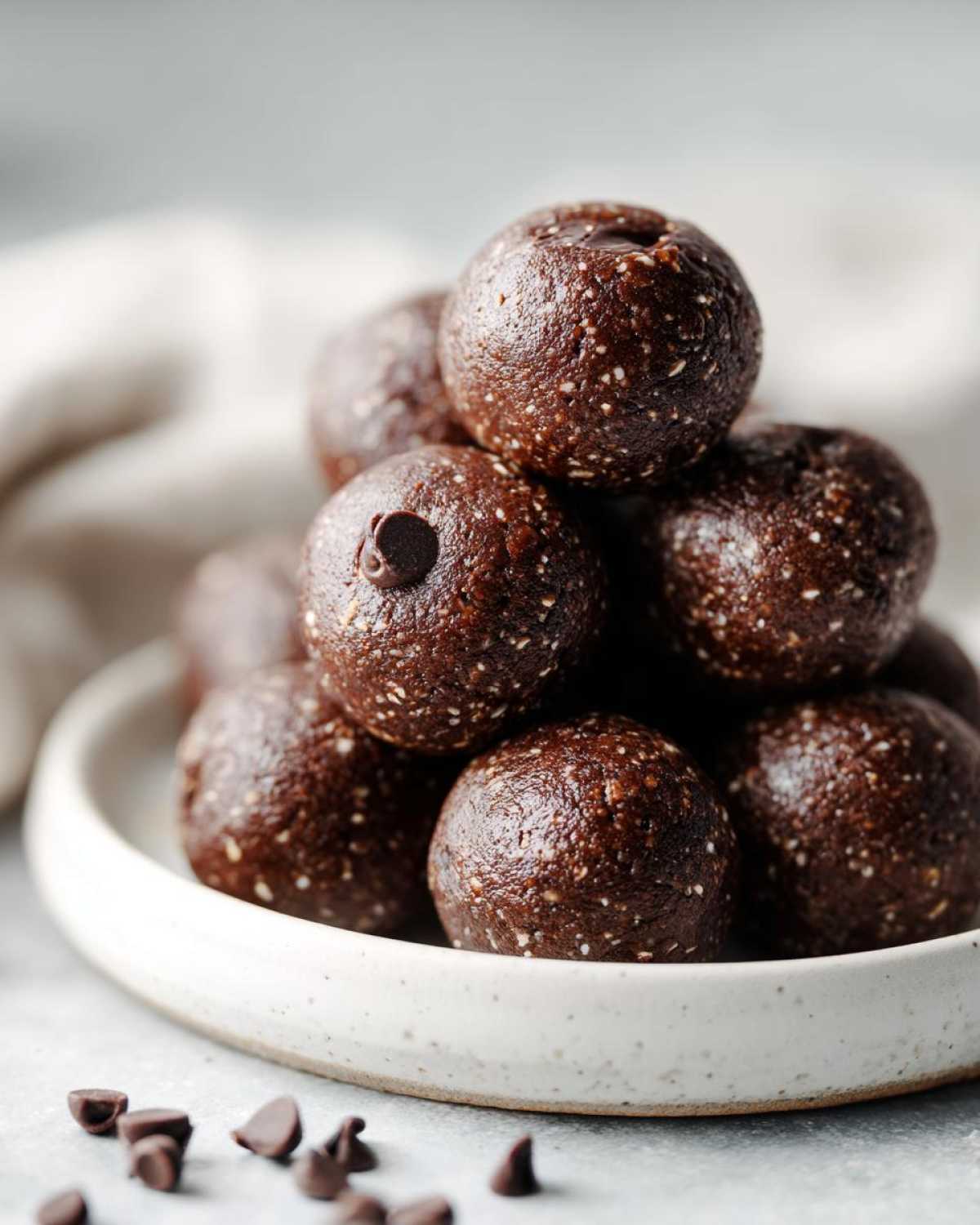 A stack of rich, dark No-Bake Brownie Batter Protein Balls topped with mini chocolate chips on a white plate.