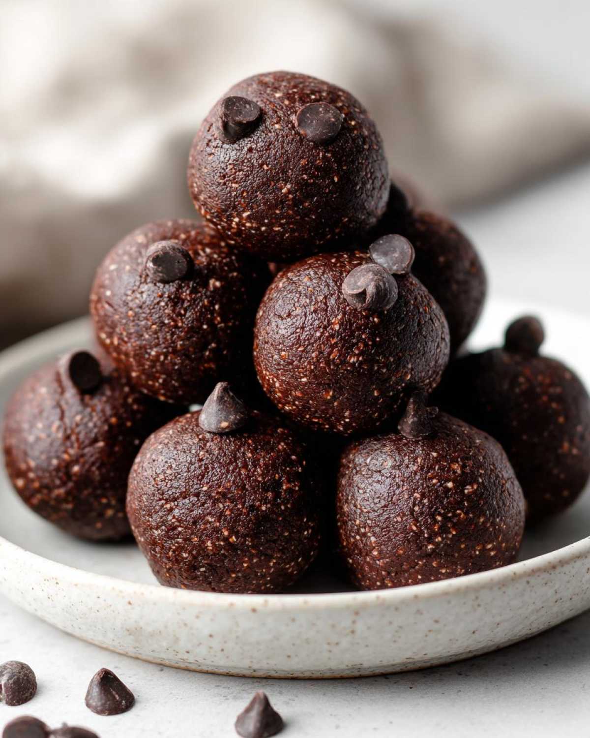 A stack of rich, dark No-Bake Brownie Batter Protein Balls topped with mini chocolate chips on a speckled white plate.