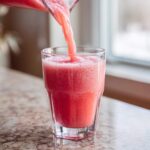 Close-up of a bright pink watermelon smoothie being poured from a pitcher into a tall glass.