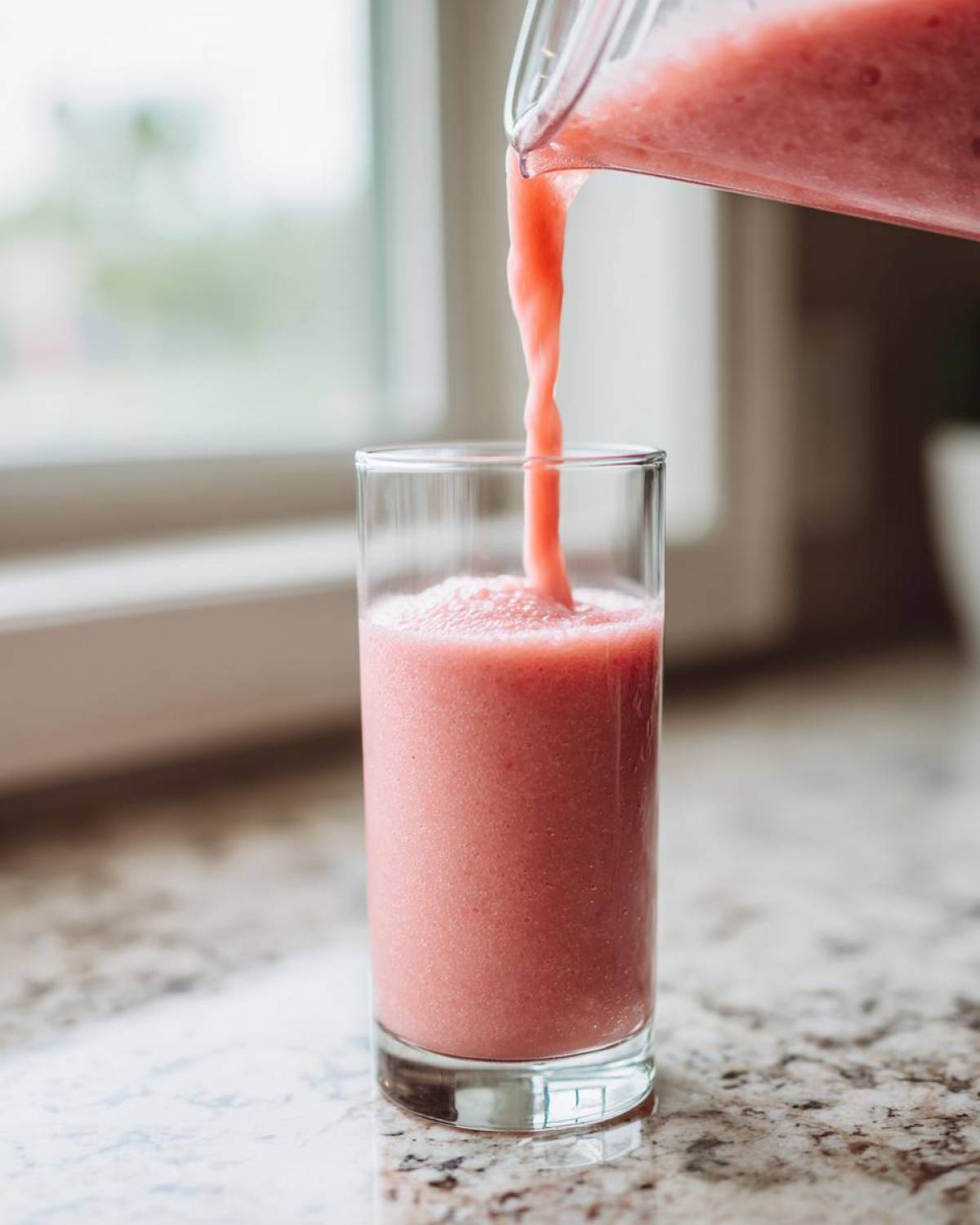 Close-up of a thick, pink watermelon smoothie being poured from a blender pitcher into a tall glass.