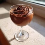 A glass of rich, dark chocolate mousse topped with shavings, sitting in sunlight.