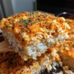 A wooden spatula lifts a thick, cheesy slice of baked sushi bake, showing layers of rice and topping.