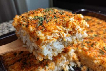A wooden spatula lifts a thick, cheesy slice of baked sushi bake, showing layers of rice and topping.
