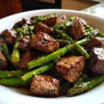 Close-up of juicy steak bites mixed with bright green asparagus, coated in garlic butter sauce from Sheet Pan Garlic Butter Steak and Asparagus.