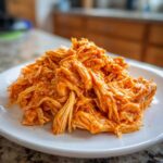 Close-up of shredded buffalo chicken coated in vibrant orange sauce piled on a white plate.