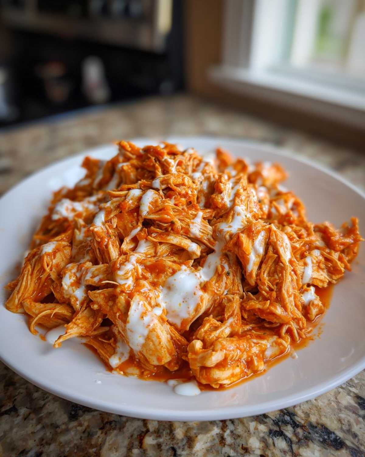Close-up of shredded buffalo chicken tossed in sauce and drizzled with ranch dressing on a white plate.