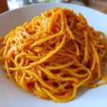 Close-up of spaghetti noodles fully coated in a vibrant orange butternut squash pasta sauce on a white plate.
