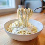 A fork lifts spaghetti noodles coated generously in Vegan Creamy Cashew Alfredo Sauce from a white bowl.