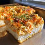 A close-up, golden-brown slice of delicious sushi bake showing a base of white rice and a creamy topping sprinkled with sesame seeds.